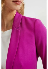 Blazer fuchsia avec un col structuré et une texture subtile, associé à un T-shirt blanc. Gros plan montrant les détails du col et de l'épaule.