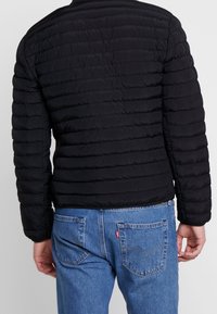 Svart puffjacka som bärs över blå jeans, sedd bakifrån, vilket visar det texturerade tyget och den övergripande avslappnade stilen.