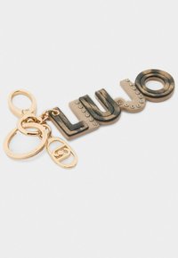 Nøglering med ordet "LUJO" i metallisk guld og sorte accenter, glatte og teksturerede bogstaver, fastgjorte ringe og dekorative nitter.