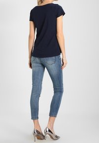 Mujer vestida con una blusa de manga corta azul navy, jeans ajustados de color azul claro y tacones altos plateados, de pie contra un fondo gris liso.