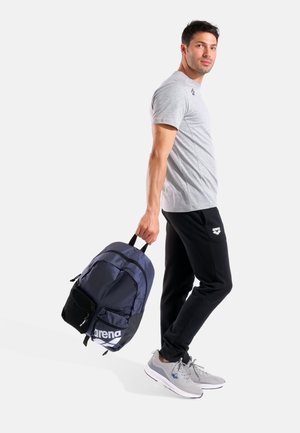 Marineblauer Rucksack mit schwarzen Akzenten, ausgestattet mit einer Netz-Tasche und einem großen Logo. Das Modell trägt ein graues T-Shirt und schwarze Hosen sowie graue Turnschuhe.