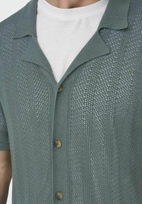 Strickjacke mit kurzen Ärmeln in gedämpftem Grün, mit einem strukturierten Muster, einem Reverskragen und braunen Knöpfen auf der Vorderseite. Getragen über einem weißen Hemd.