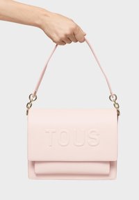 Rosa Lederhandtasche mit geprägtem "TOUS"-Logo, flachem Design und doppeltem Tragegriff aus goldfarbenem Material zur Befestigung.