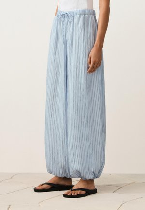 Person wearing light blue pinstriped balloon pants with elastic cuffs and a drawstring waist, paired with black flip-flops on a tile floor.