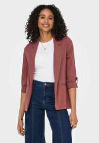  Rostfarbener Blazer mit hochgerollten Ärmeln und Revers, getragen über einem weißen T-Shirt, kombiniert mit dunklen Jeans. Glatte Stoffoberfläche.