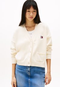 Kremfarget cardigan med v-hals, knappeåpning og lommer. Har ribbestrikkede mansjetter og kant; kombinert med et denimskjørt.