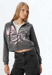 Sudadera gris recortada con estampados gráficos en rosa y texto blanco, con cremallera frontal y capucha con cordón. Combinada con pantalones cargo oscuros.