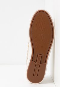 Semelle d'une basket beige avec une bande de caoutchouc marron, présentant une surface texturée antidérapante et le nom « MICHAEL KORS » embossé sur la zone du talon.