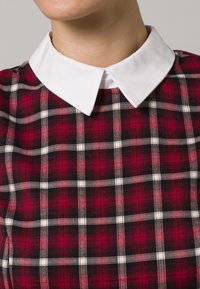 Červený a černý károvaný vzor s bílými akcenty; obsahuje klasický bílý límeček. Materiál vypadá texturovaně a tkaně.