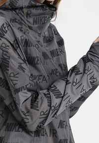 Veste grise avec un imprimé noir "NIKE AIR" sur l'ensemble, un col montant, une fermeture éclair à l'avant et des manches ajustées. Le matériau semble léger et texturé.