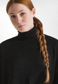 Schwarzer, weicher Rollkragenpullover mit strukturierter Oberfläche, lässiger Schnitt. Lange, geflochtene, kastanienbraune Haare fallen über eine Schulter.