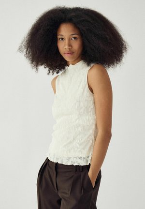 Ung kvinde med voluminøst krøllet hår iført en ærmeløs hvid tekstureret top og mørke bukser med høj talje, stående mod en ensfarvet baggrund.