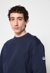 Sweatshirt bleu marine à col rond en tissu texturé avec une petite écusson rond sur la manche gauche et des détails de couture visibles.