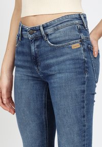 Nahaufnahme eines Oberkörpers, der blaue Jeans mit einem kleinen braunen Etikett an der rechten Tasche und einem kurzen cremefarbenen Strickoberteil trägt.