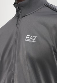 Chaqueta deportiva gris con cremallera, hecha de una tela suave, que presenta un cuello alto y el logo de EA7 Emporio Armani en el lado izquierdo del pecho.