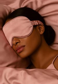 Vrouw met donker haar die een roze satijnen slaapmasker en bijpassende lakens draagt, liggend met bedekte ogen en een ontspannen uitdrukking.