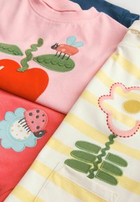 Tres camisetas de algodón: rosa con un corazón rojo y un insecto, coral con un diseño de flores, y crema a rayas con bordados de flores y hojas.