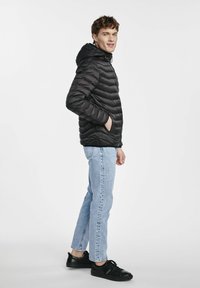 Czarna kurtka puchowa z poziomym pikowaniem, kapturem i zamykanymi kieszeniami, noszona z jasnymi dżinsami i czarnymi trampkami. Casualowy styl.