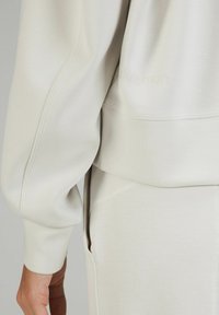 Primer plano de la mano de una persona descansando a su lado, vestida con una camiseta de manga larga de color beige claro y pantalones a juego con detalles de costura visibles.
