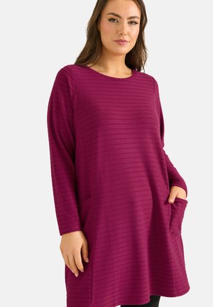 Mujer con un vestido túnica púrpura de manga larga y textura, con bolsillos, una mano en el bolsillo, de pie contra un fondo claro y liso.