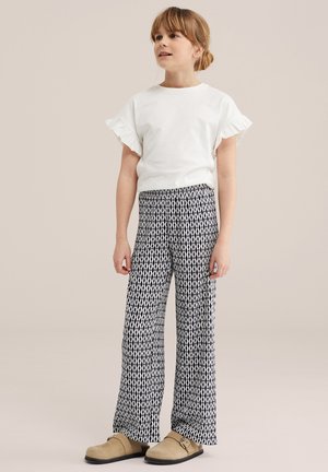 Fată cu păr castaniu deschis poartă un top alb cu mâneci scurte cu volane, pantaloni largi cu model alb-negru și pantofi slip-on bej.