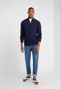 Pullover de malha azul marinho com um fecho de correr até um quarto, combinado com jeans azul claro. Ténis coloridos com detalhes em verde, amarelo e vermelho.