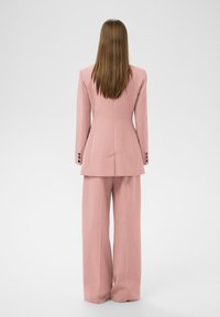 Rosa maßgeschneiderter Anzug mit einem taillierten Blazer, der strukturierte Schultern und weitgeschnittene Hosen umfasst. Glatter Stoff mit Knopfakzenten an den Ärmeln.