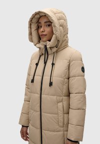 Bežinė pūkinė striukė su didele, išklota kapuce, priekyje su užtrauktuku, raišteliais, šoninėmis kišenėmis ir apvalia logotipo etikete ant rankovės.