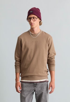 Jonge man met een bril, bordeauxrode muts, beige sweatshirt, gelaagd overhemd, grijze spijkerbroek en een zilveren ketting tegen een effen lichte achtergrond.