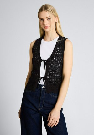 Kobieta nosząca czarną, bezrękawnikową kamizelkę na szydełku, związaną z przodu, na białym topie i ciemnoniebieskich dżinsach, stojąca na jednolitym tle.