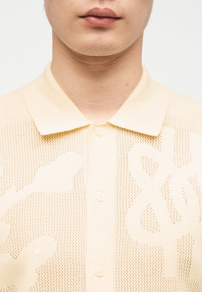 Κοντινό πλάνο ενός ατόμου που φοράει ένα κρεμ πλεκτό πόλο πουκαμίσα με κουμπιά, διακριτικό υφή και γιακά.