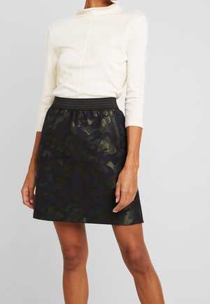 Frau trägt ein weißes, eng anliegendes Langarmshirt und einen schwarzen Rock mit Camouflage-Muster und elastischem Bund, steht vor einem einfarbigen Hintergrund.