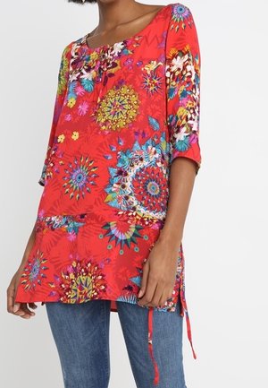 Femme portant une tunique rouge avec des motifs floraux et géométriques colorés et des manches trois-quarts, associée à un jean bleu.