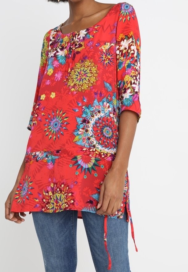 Femme portant une tunique rouge avec des motifs floraux et géométriques colorés et des manches trois-quarts, associée à un jean bleu.
