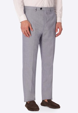 Uomo che indossa pantaloni formali grigio chiaro, camicia bianca e scarpe slip-on marrone scuro, in piedi con le mani rilassate lungo i fianchi.