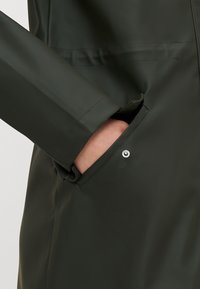 Zielona wodoodporna kurtka o eleganckim designie, z boczną kieszenią zamykaną na srebrny zatrzask oraz gładką teksturą.
