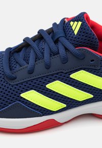 Zapatilla deportiva de color azul marino con parte superior de malla, rayas en verde neón y acentos rojos. Presenta materiales texturizados y un diseño tradicional de cordones.