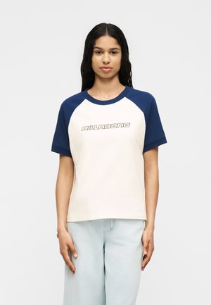 Γυναίκα με μακριά σκούρα μαλλιά, φορώντας λευκό και ναυτικό μπλουζάκι Billabong με raglan μανίκια και ανοιχτό μπλε παντελόνι, στέκεται μπροστά σε απλό φόντο.