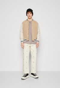 Jakna bomber v dveh tonih, tan in smetana, s črno-belim pr trimom, siva majica, off-white hlače z majhnim vzorcem pokalov, črne superge.