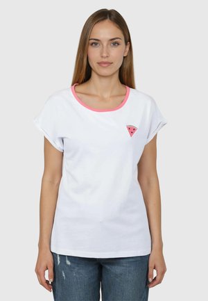 Kobieta z prostymi brązowymi włosami nosi białą koszulkę z różowym dekoltem i małą naszywką przedstawiającą plaster arbuza na lewej piersi, w połączeniu z niebieskimi dżinsami.