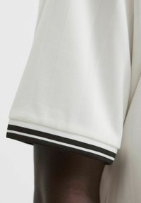 Camisa blanca de manga corta con puños acanalados a rayas blancas y negras, sobre un brazo de piel oscura, contra un fondo claro.