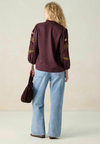 Maroen blouse met geplooide mouwen en bloemrijke borduursels. Gecombineerd met lichtblauwe jeans met wijde pijpen en een kleine bordeauxrode handtas.