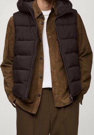 Mężczyzna w czarnej pikowanej kamizelce bez rękawów na brązowej koszuli na guziki i brązowych spodniach, stoi z rękami w kieszeniach.