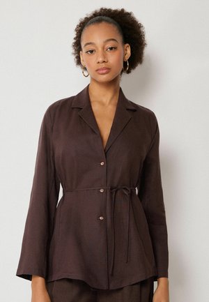 Frau mit natürlichen lockigen Haaren trägt eine braune Hemdjacke mit Knöpfen und Bindegürtel, steht vor einem schlichten hellen Hintergrund.