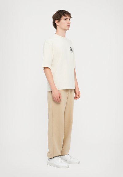 T-shirt crème à manches courtes avec un design graphique sur la poitrine gauche, associé à un pantalon beige ample et des baskets blanches.