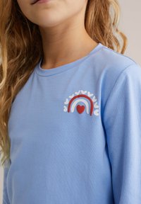 Camisa de manga larga azul claro con un diseño de arcoíris con corazones, detallada con acentos en rojo, blanco y rosa. Textura suave.