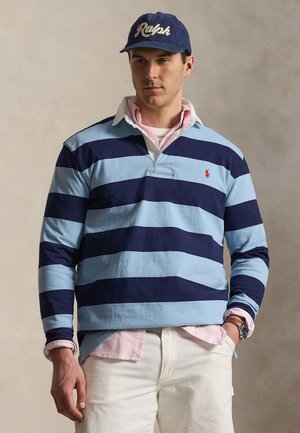 Mann trägt ein marineblau und hellblau gestreiftes Rugby-Shirt, weiße Hose, rosa Hemd mit Kragen darunter, marineblaue Kappe mit dem Schriftzug „Ralph“ und Armbanduhr.