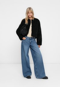 Ανοιχτόχρωμη γυναίκα που φοράει μια μαύρη φuzzy τζάκετ, μπεζ τοπ, μπλε τζιν με φαρδιά πόδια, μαύρα παπούτσια και κρατάει μια μαύρη τσάντα ώμου.