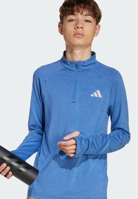 Modrý športový pulóver s štvrtinovým zipsom a dlhými rukávmi. Obsahuje štruktúrovanú tkaninu a biele logo na hrudi. Drží čiernu fľašu na vodu.