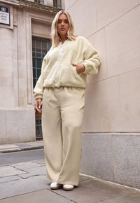 Kremowa, puszysta kurtka na zamek błyskawiczny z bocznymi kieszeniami; zestawiona z szerokimi, beżowymi spodniami. Obuwie to białe, zaokrąglone, niskie buty na obcasie.